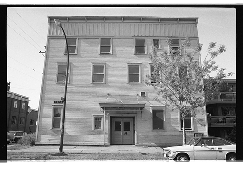 Black and white photograph of Western Front's three-storey wood-clad building on a street corner, with a street sign reading "E 8th" visible at the lower left. The building has a flat roofline, double entry doors at street level, and several rectangular windows. A young tree stands to the right of the entrance, and vintage cars are parked on the street. A neighbouring apartment building is partially visible on the right.