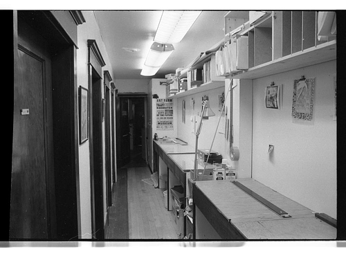 A black and white photography of a narrow hallway with a run of five dark wooden doors on the lefthand side and a row of workbenches and shelves hung close to the ceiling on the right hand side. A number of posters, drawings and photographs can be seen pinned to the wall, while paper files, folders, and slide carousels and boxes can be seen on the bench tops and shelves. The hallway is brightly lit from above by fluorescent lights.
