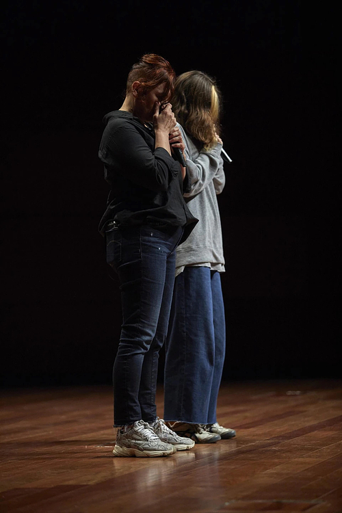 Two people stand close together on a wooden stage, each holding a microphone. The person in front, dressed in a black shirt and jeans, has their eyes closed and one hand raised to their face as if wiping away tears. The person behind them, wearing a grey sweatshirt and wide blue pants, faces the same direction with their head slightly bowed. The lighting is dim, creating an intimate atmosphere.