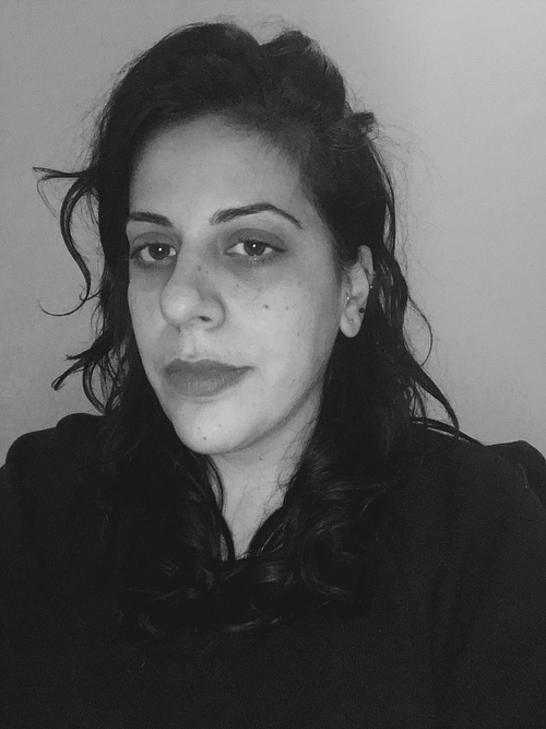 Black-and-white portrait of Suzy Halajian, a woman with dark curly hair worn loose over her shoulders and gathered at the top, looking directly at the camera with a neutral expression. She wears a dark, high-necked top and small stud earrings, photographed against a plain light-gray background.