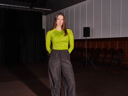 Nina Davies stands in theatrical space. She looks directly at the camera with her hair down and hands placed behind her back. She wears black tracksuit pants and a bright lime coloured sports top.  