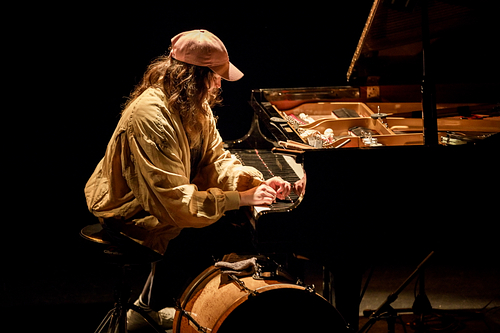 Eve Risser wears a loose beige jacket and a pink baseball cap while playing a grand piano on a dimly lit stage. She is seated on a drum stool, with a large bass drum positioned below the keyboard. The piano's lid is open, revealing various objects placed inside the instrument, suggesting a prepared piano setup. The scene is intimate and atmospheric, with warm lighting highlighting the performer’s focused posture.